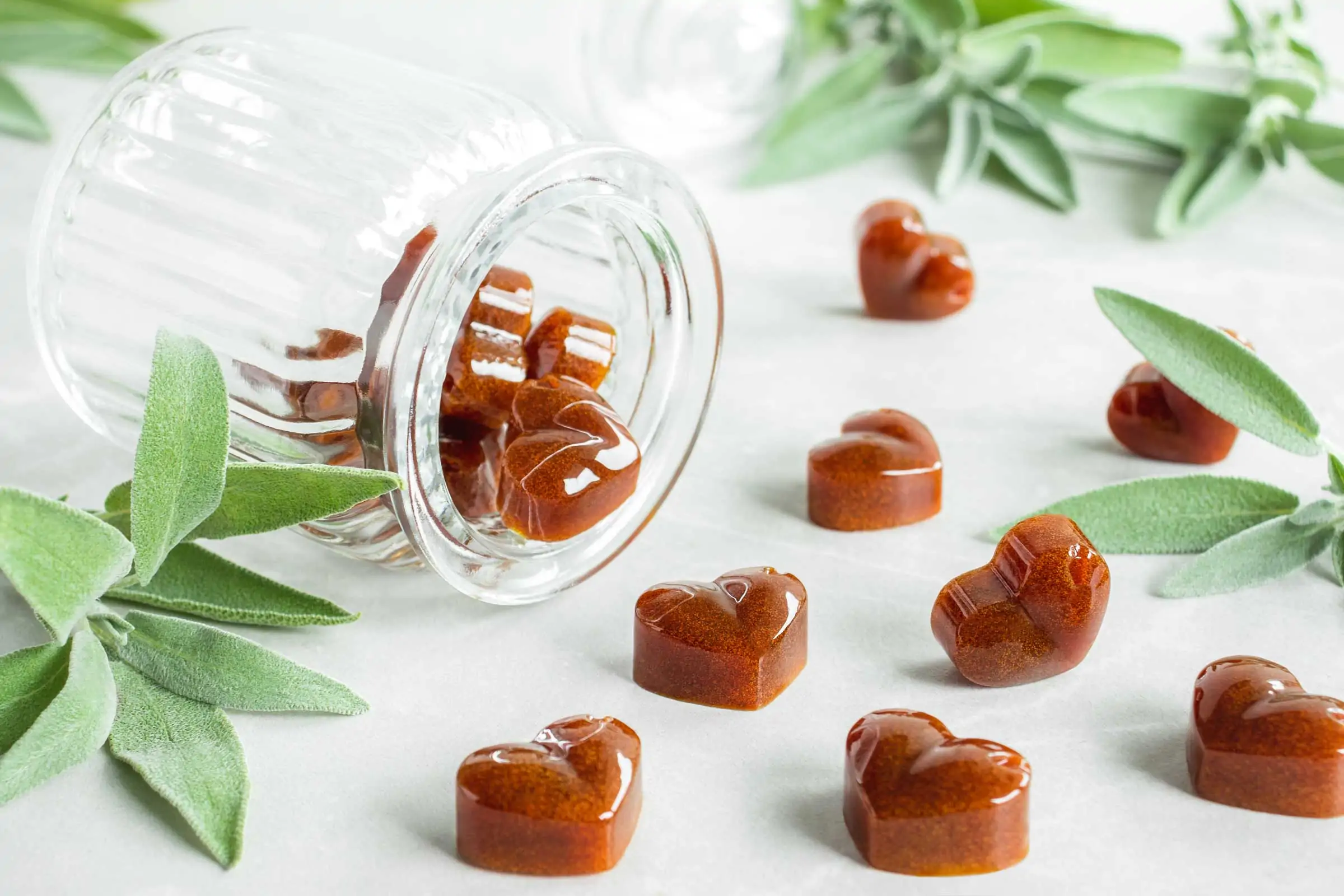 Salbeibonbons in Herzform liegen neben einem Bonbonglas und frischem Salbei.