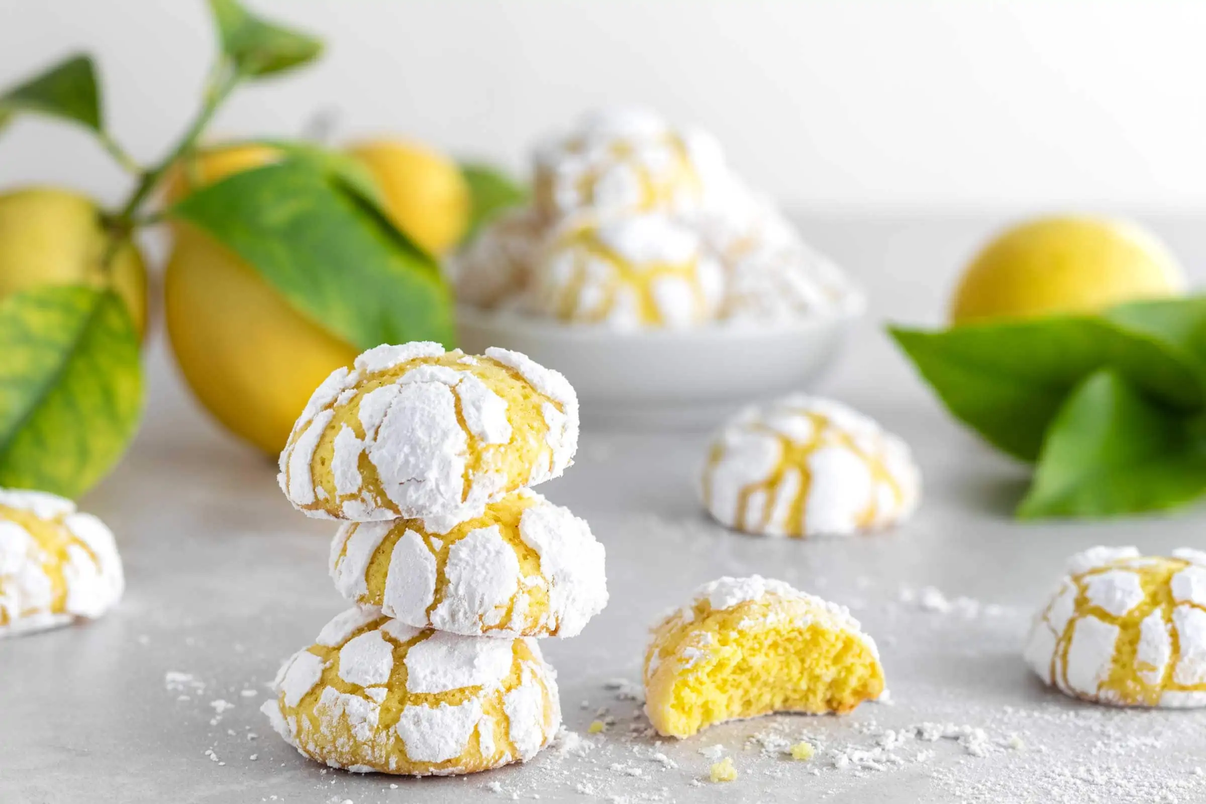 Lemon Crinkle Cookies aufeinander gestapelt mit frischen Zitronen im Hintergrund. Eines der Zitronenplätzchen daneben ist angebissen.
