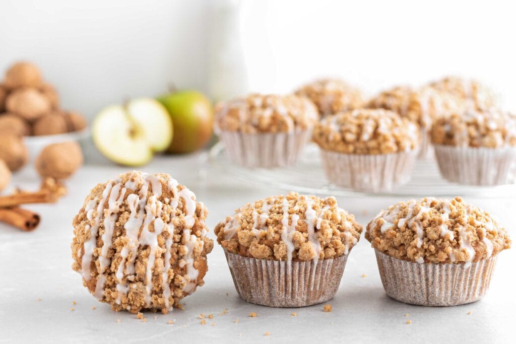 Saftige Apfel-Walnuss-Muffins mit Zimtstreuseln aufgereiht. Im Hintergrund stehen weitere Muffins auf einem Abkühlgitter.