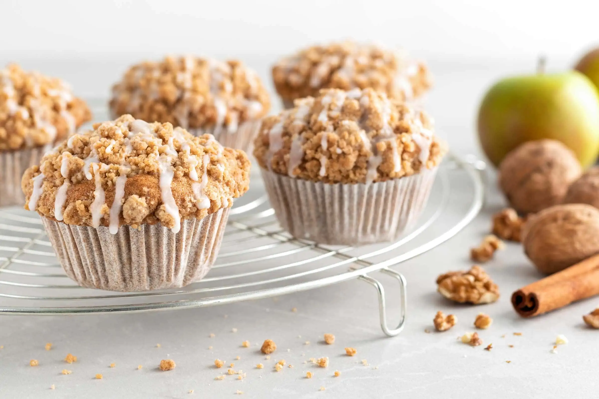 Apfel-Walnuss-Muffins auf einem Abkühlgitter. Daneben liegen Walnüsse, Zimtstangen und ein Apfel.