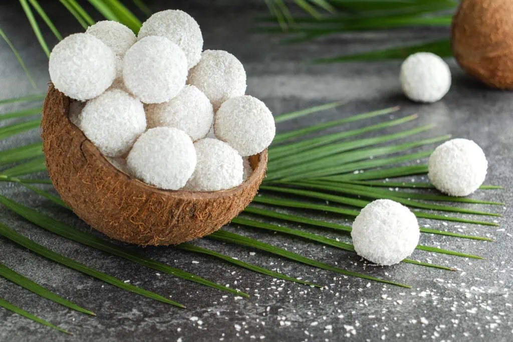 Kokospralinen umhüllt mit weißen Kokosraspeln liegen in einer Schale aus einer Kokosnuss. Unter der Schale mit den Pralinen liegt ein Palmenblatt.