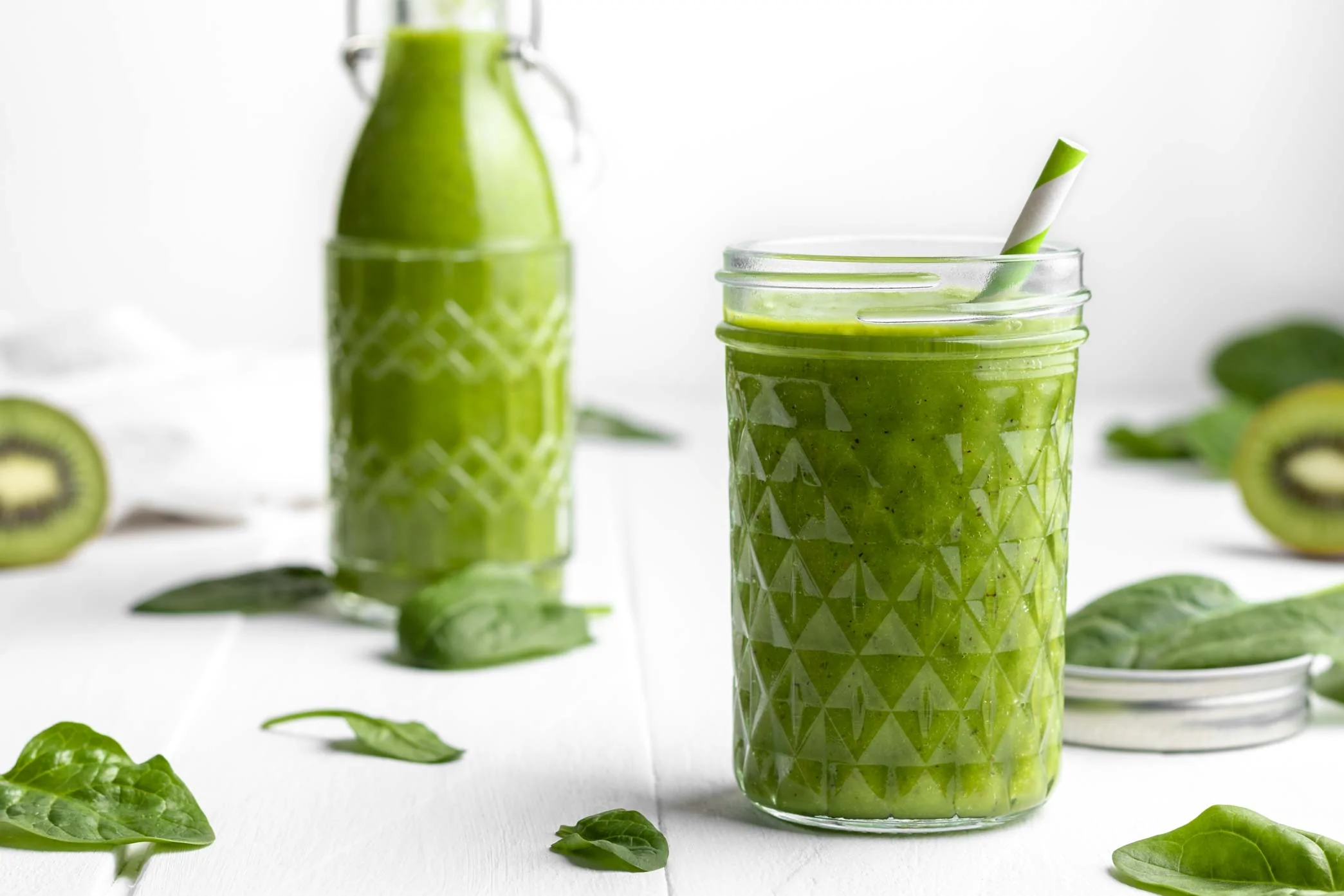 Gesunder grüner Smoothie mit Spinat, Banane und Mango in einem Glas mit Strohhalm. Neben dem Glas liegen Spinatblätter und halbierte Kiwis.