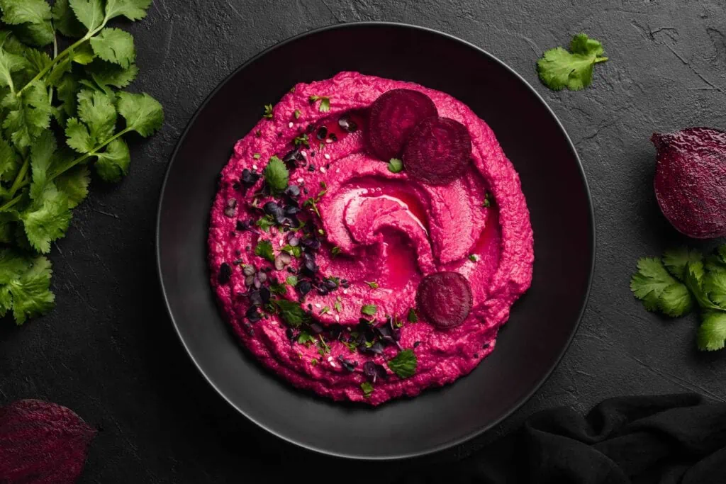 Rote Bete Hummus in einem Teller garniert mit Rote Bete Scheiben, Sesam, Koriander und Kresse.