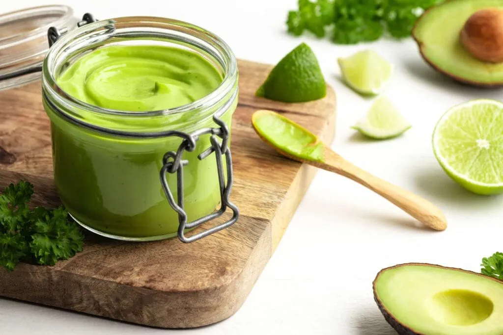 Avocado-Limetten-Mayonnaise ohne Ei in einem Glas. Daneben liegen geschnittene Avocados und Limetten.
