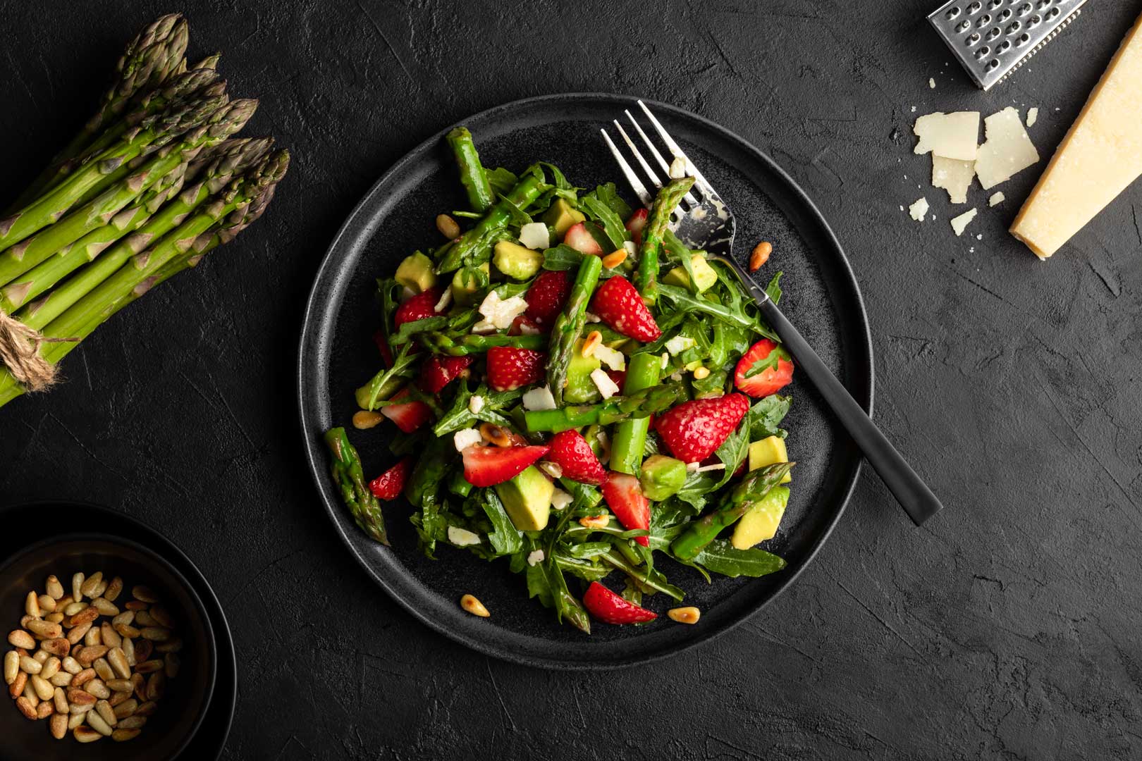 Grüner Spargel-Salat mit Erdbeeren und Avocado auf einem schwarzen Teller. Neben dem Salat liegen grüner Spargel, Parmesan und Pinienkerne.