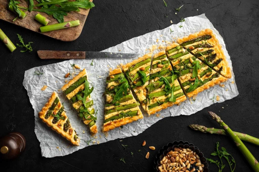 Feine grüne Spargeltarte mit Blätterteig in Stücke geschnitten auf einem Backpapier. Auf der Tarte liegen Pinienkerne und Rucola.