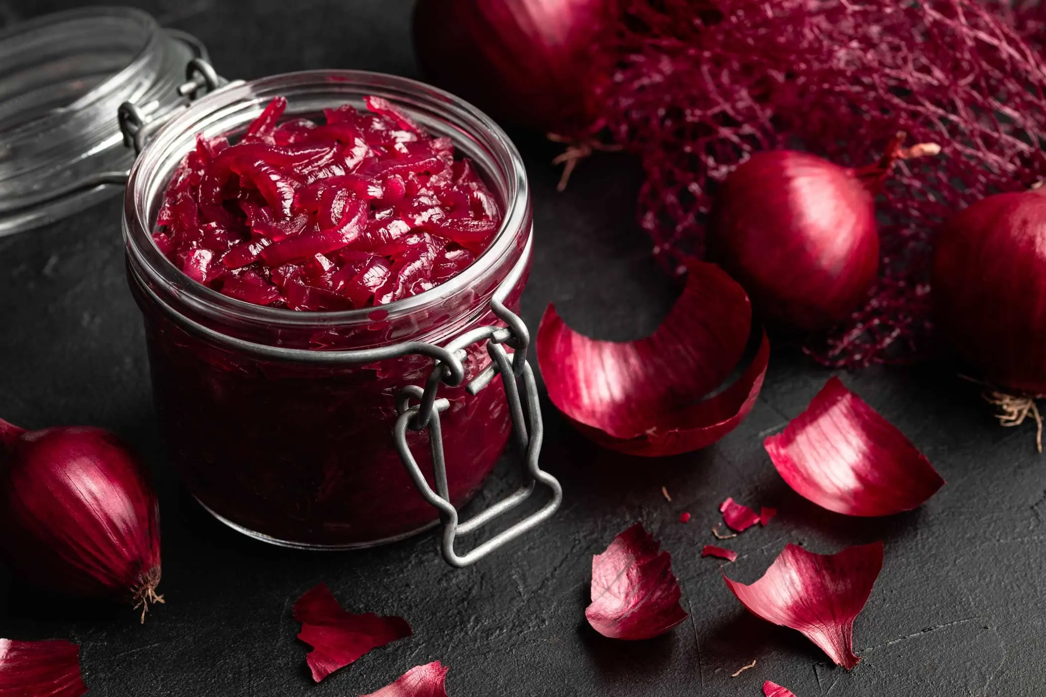 Rotes Zwiebel Chutney in einem Bügelverschlussglas neben einem Netz roter Zwiebeln.