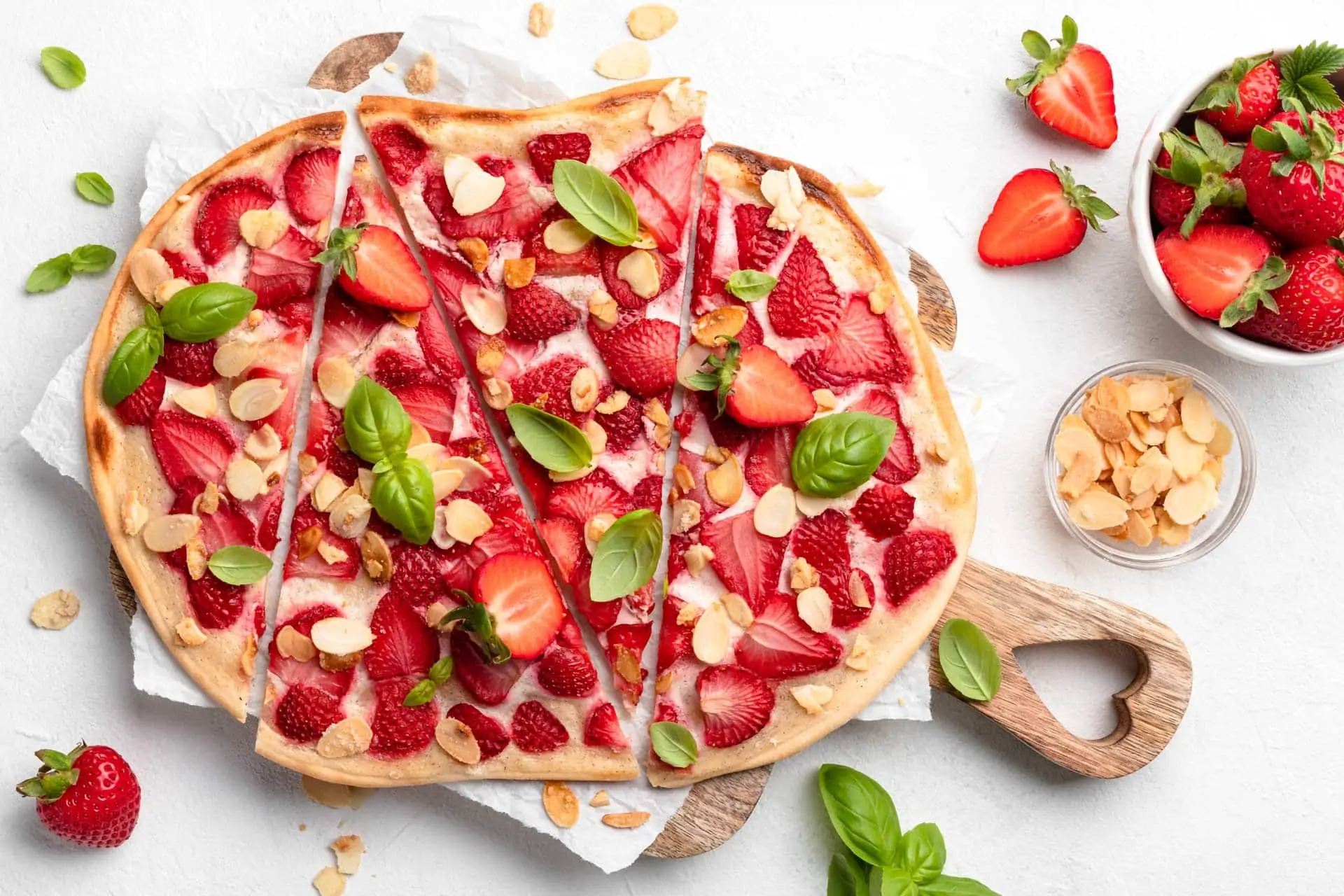 Süßer Flammkuchen mit Erdbeeren, garniert mit karamellisierten Mandeln und frischen Basilikumblättern, auf einem Holzbrett.
