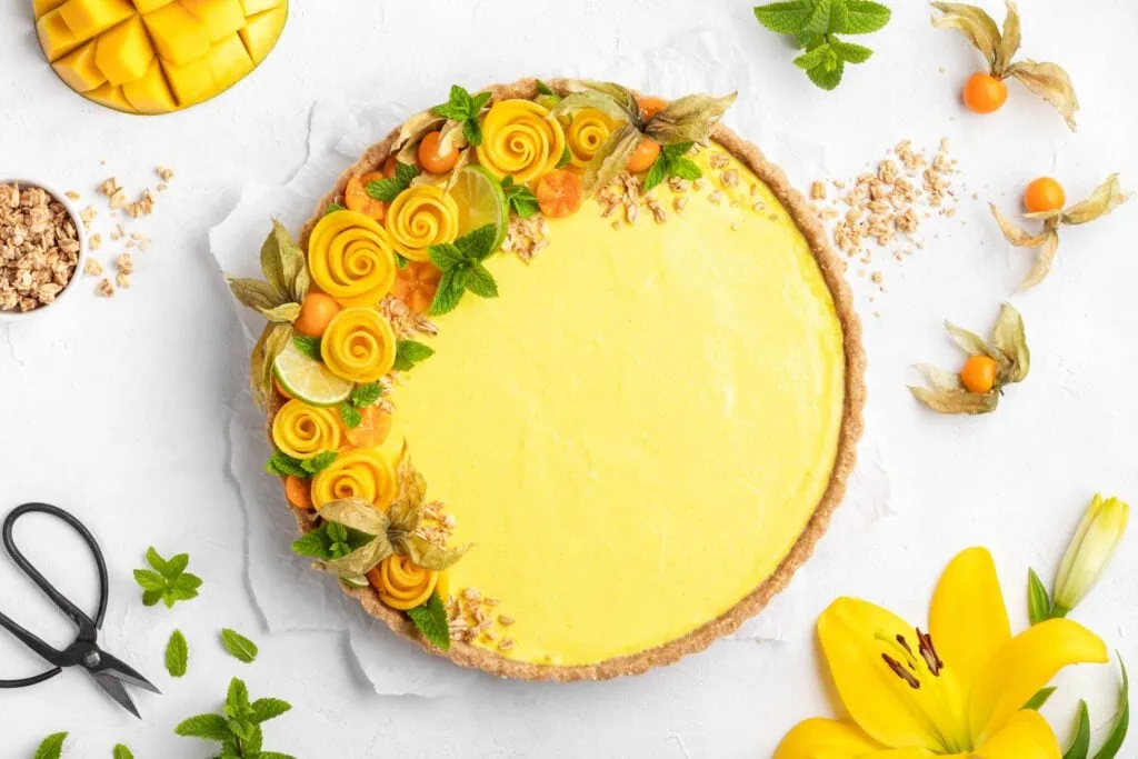 No Bake Mango Tarte garniert mit Granola, Mangorosen, Minze und Physalis auf einem weißen Untergrund.