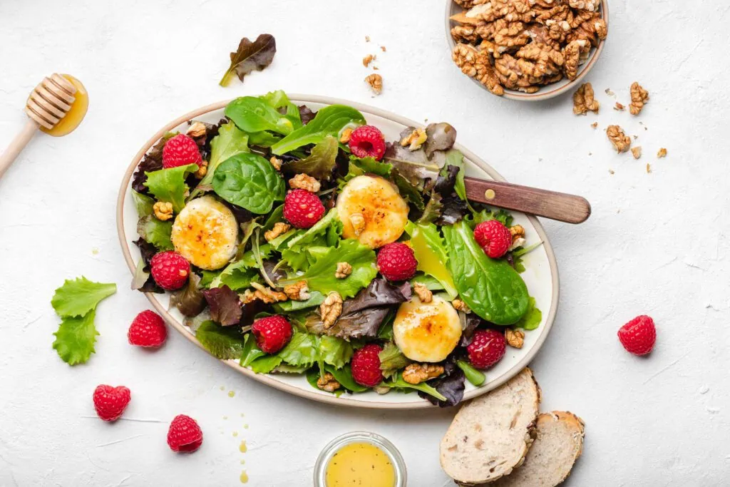 Salat mit gebackenem Ziegenkäse mit Honig und Walnüssen und frischen Himbeeren auf einem ovalen Teller.
