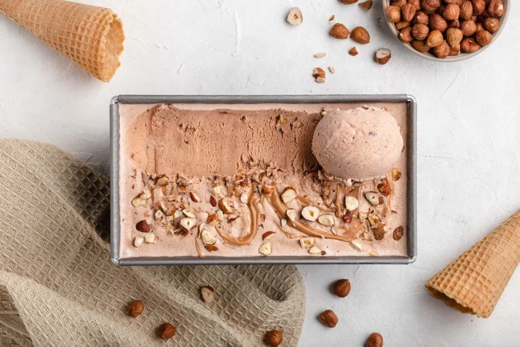 Neben der Form gefüllt mit dem cremigen Haselnusseis liegen Eiswaffeln und ein Schälchen mit gerösteten Haselnüssen.