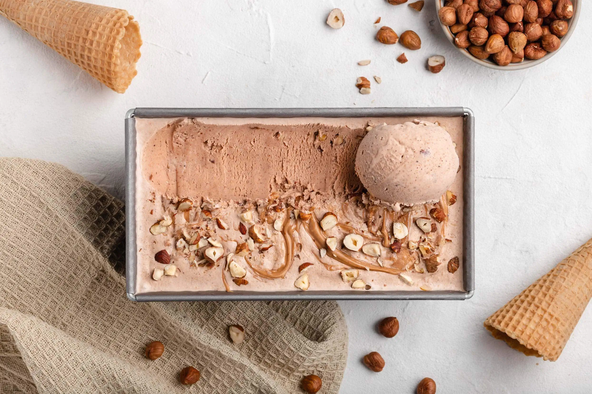 Neben der Form gefüllt mit dem cremigen Haselnusseis liegen Eiswaffeln und ein Schälchen mit gerösteten Haselnüssen.
