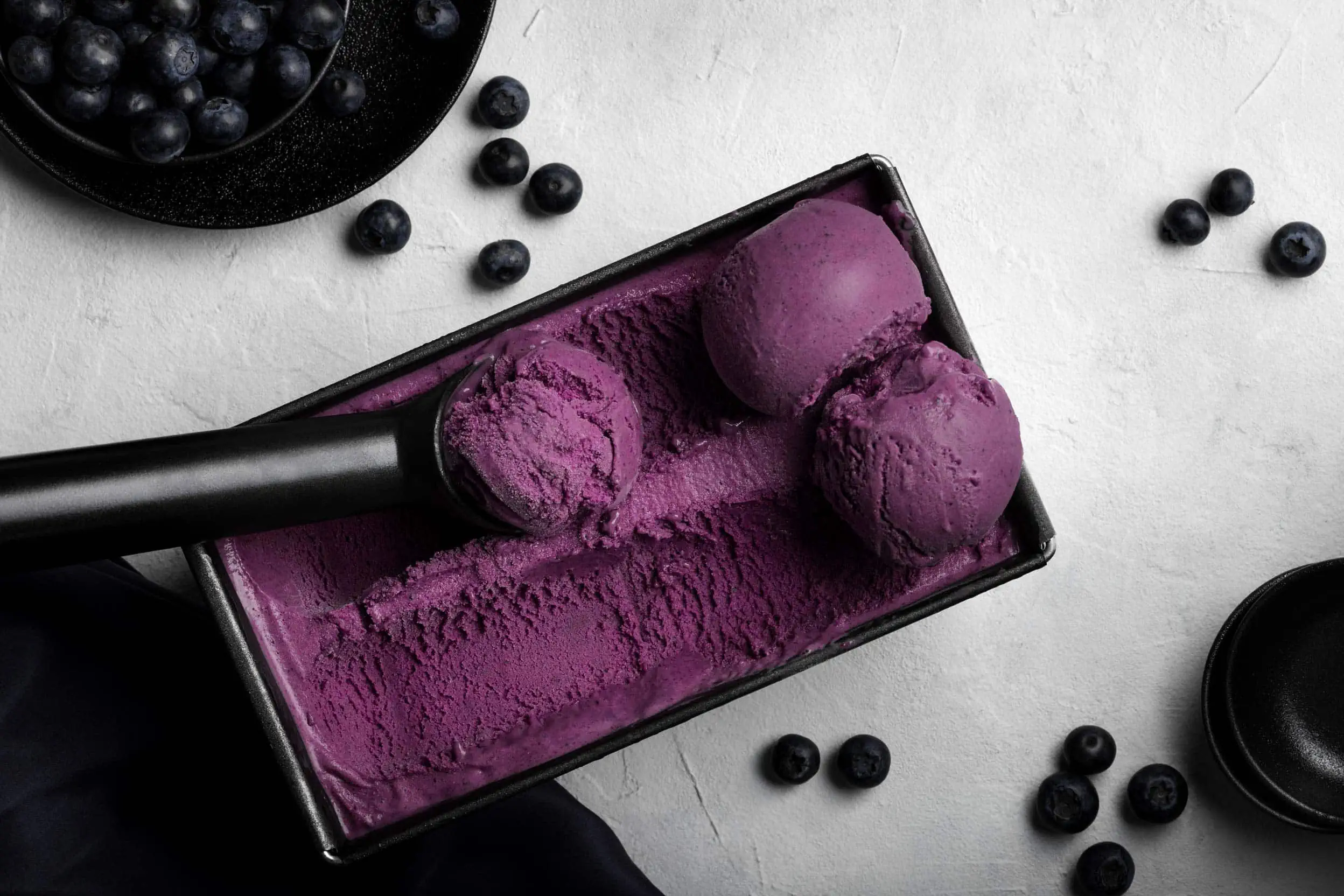 Das fertig zubereitete, fruchtige Blaubeereis aus der Eismaschine in einer Form. Daneben liegen frische Blaubeeren.