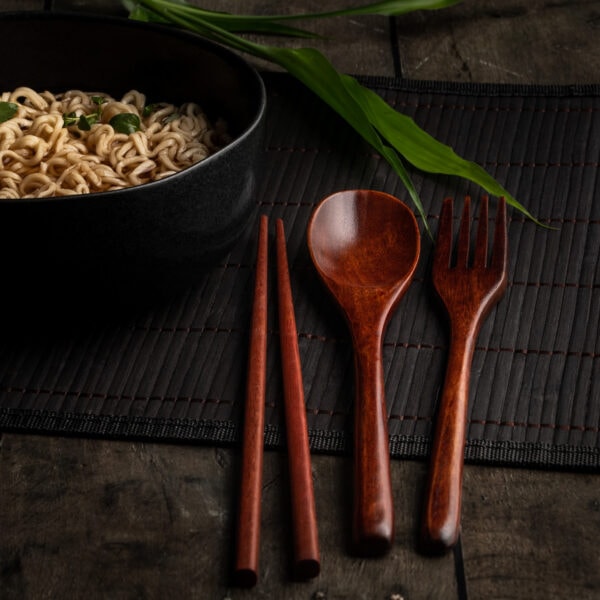 Ein asiatisches Besteck aus Holz auf einem Tischset aus Bambus neben einer Schüssel mit Nudeln.