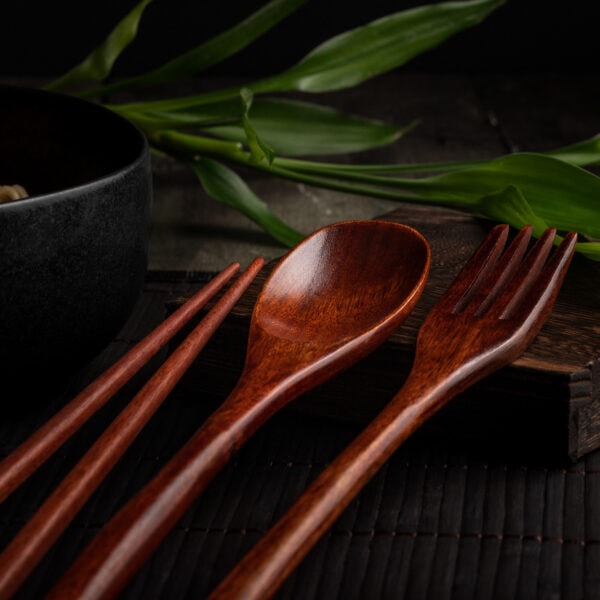 Ein handgefertigtes, asiatisches Besteck-Set aus Holz auf einem dunklen Untergrund. Das Set besteht aus Essstäbchen, einem Löffel und einer Gabel.