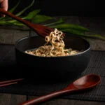 Aus einer Schüssel werden Nudeln mit einer Holzgabel aus dem asiatischen Besteck-Set herausgenommen.