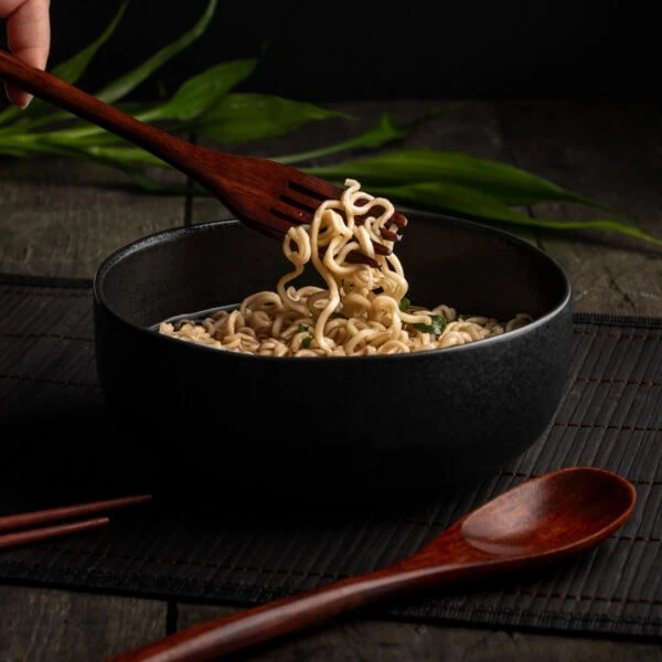 Aus einer Schüssel werden Nudeln mit einer Holzgabel aus dem asiatischen Besteck-Set herausgenommen.