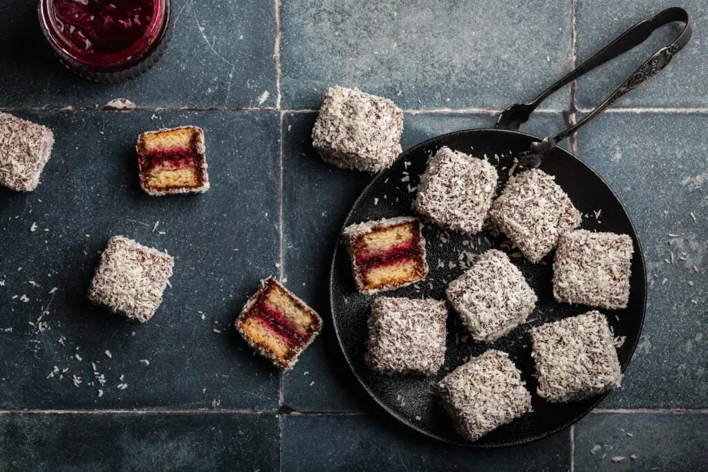 Lamingtons, ein australisches Kaffeegebäck, aus Biskuit, einer Schokoladenglasur, Kokosflocken und einer Marmeladenfüllung auf einem Teller.