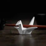 Ein weißer Origami-Kranich vor einer kleinen Schüssel auf dem ein paar Essstäbchen liegen.