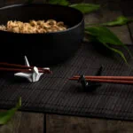 Zwei Essstäbchenhalter in Form von Kranichen vor einen Schüssel mit Nudeln auf denen Stäbchen aufliegen.