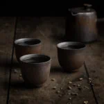 Eine Kyusu-Teekanne vor der drei asiatische Teetassen gefüllt mit grünem Tee stehen.