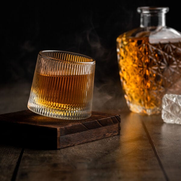 Ein Wackelglas gefüllt mit Whisky auf einem Holzuntersetzer. Dahinter steht eine Flasche mit Whisky.
