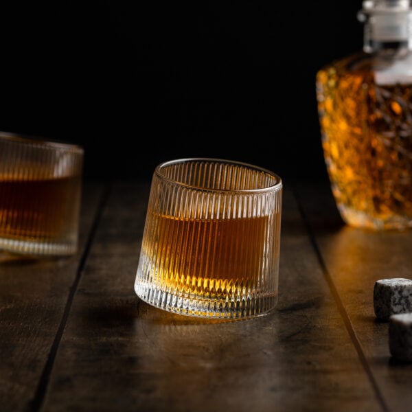 Zwei Wackelgläser mit rundem Boden stehen auf einem Holztisch. Hinter den Gläsern steht eine Karaffe mit Whisky.