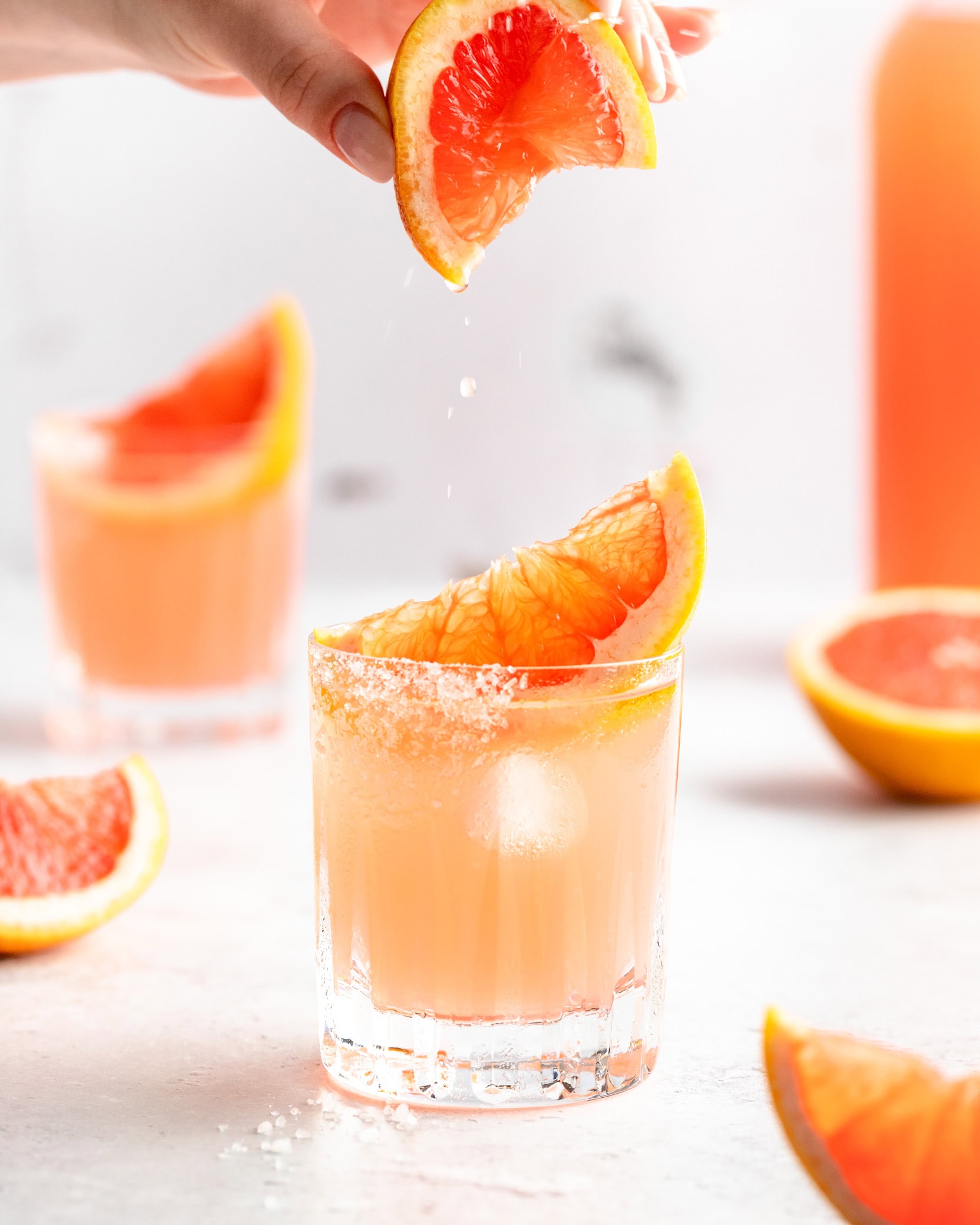 Grapefruitscheibe wird über einem Tumbler mit Pink Paloma ausgepresst. Garniert ist der mexikanische Tequila-Cocktail klassisch mit einem Salzrand.