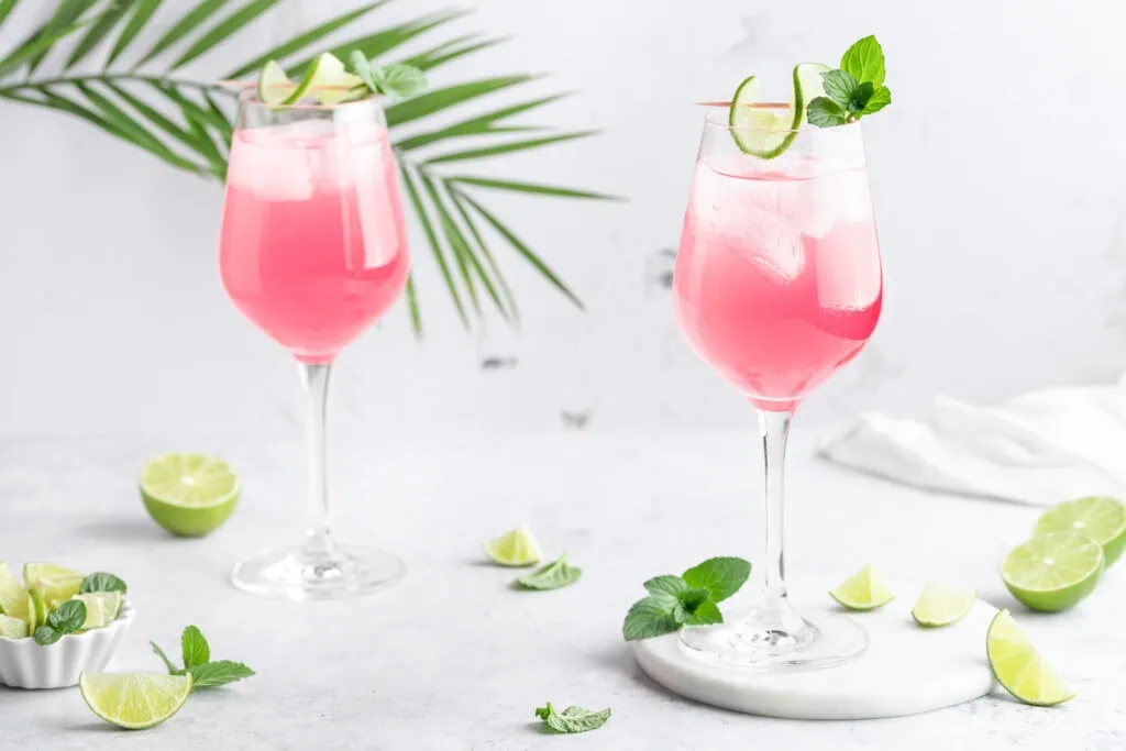Zwei rosafarbene Sarti Spritz in Weingläsern, garniert sind die Aperitifs einer Limettenscheibe.