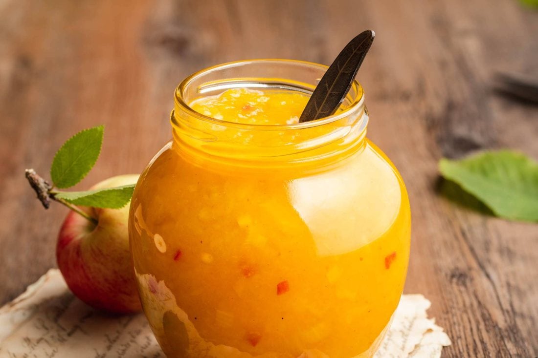 Gelb-orangenes Apfel Chutney mit Chili in einem geöffneten Schraubglas mit einem Löffel darin.
