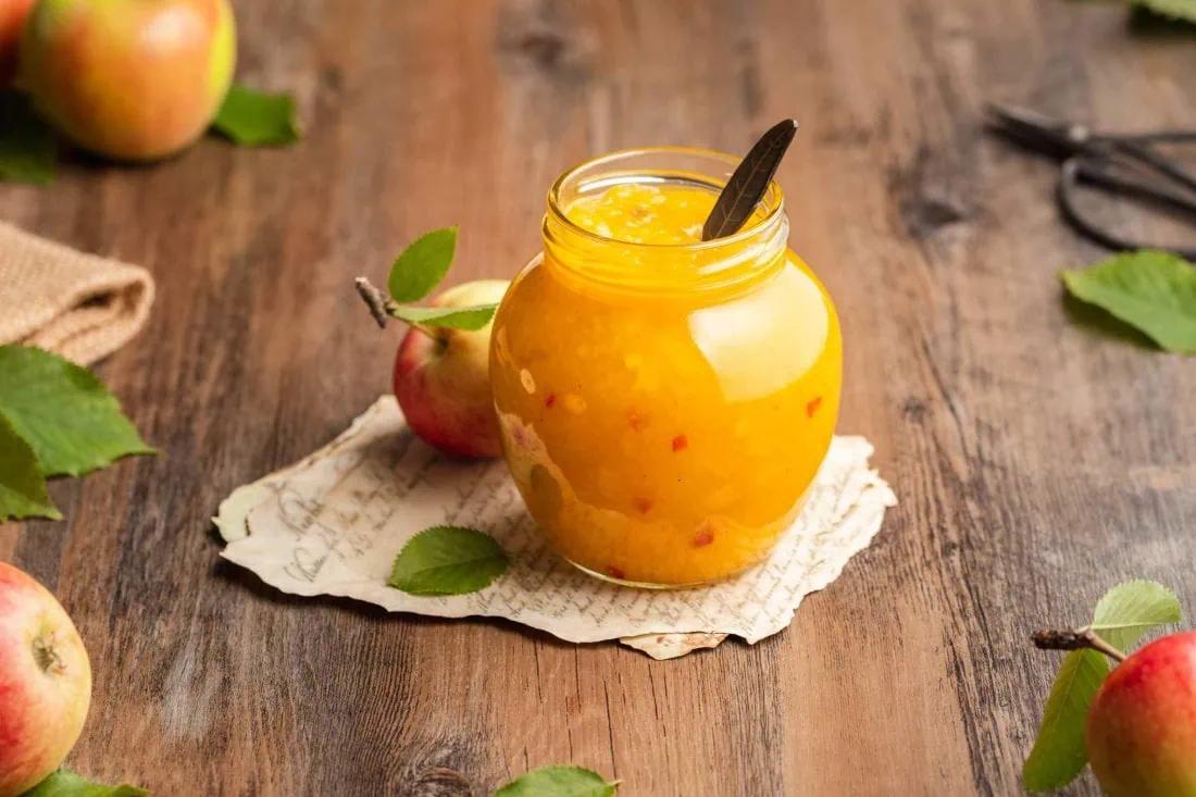 Indisches Apfel Chutney mit Chilistücken in einem Glas mit einem Löffel darin. Neben dem Glas liegen Blätter, Äpfel und eine Schere.