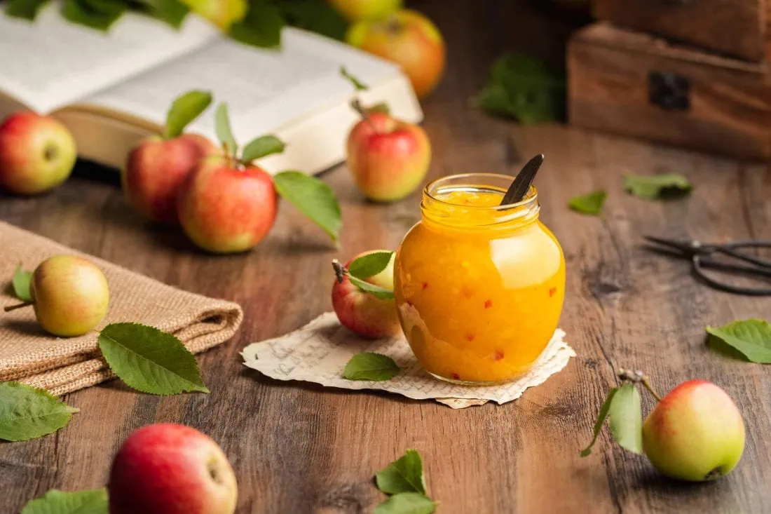 Apfel Chutney mit kleinen Chilistücken in einem runden Schraubglas. Neben dem Chutney liegen frische Äpfel und Blätter.