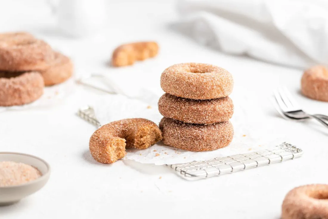 Ein Stapel Apfel-Donuts aus dem Backofen auf einem Abkühlgitter. Neben den Donuts steht ein Schälchen mit Zimt und Zucker.
