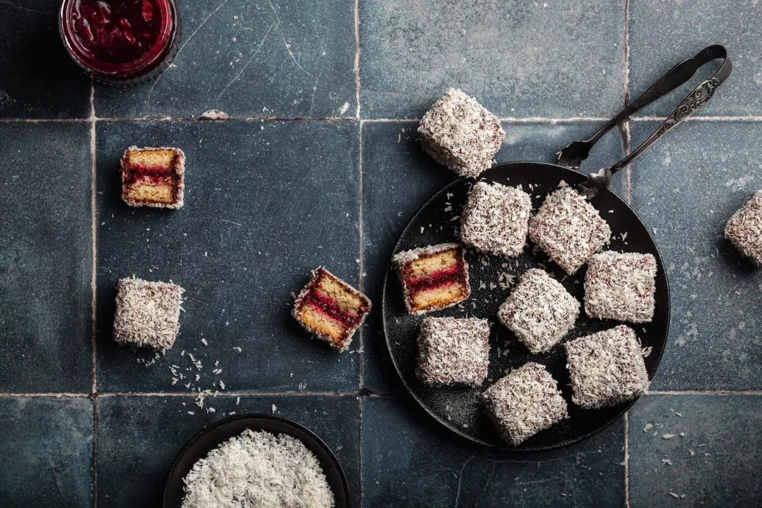 Mehrere australische Lamingtons, Kuchen mit Kokos, einer Schokoladenglasur und Marmelade, liegen zum Teil halbiert auf einem Teller.