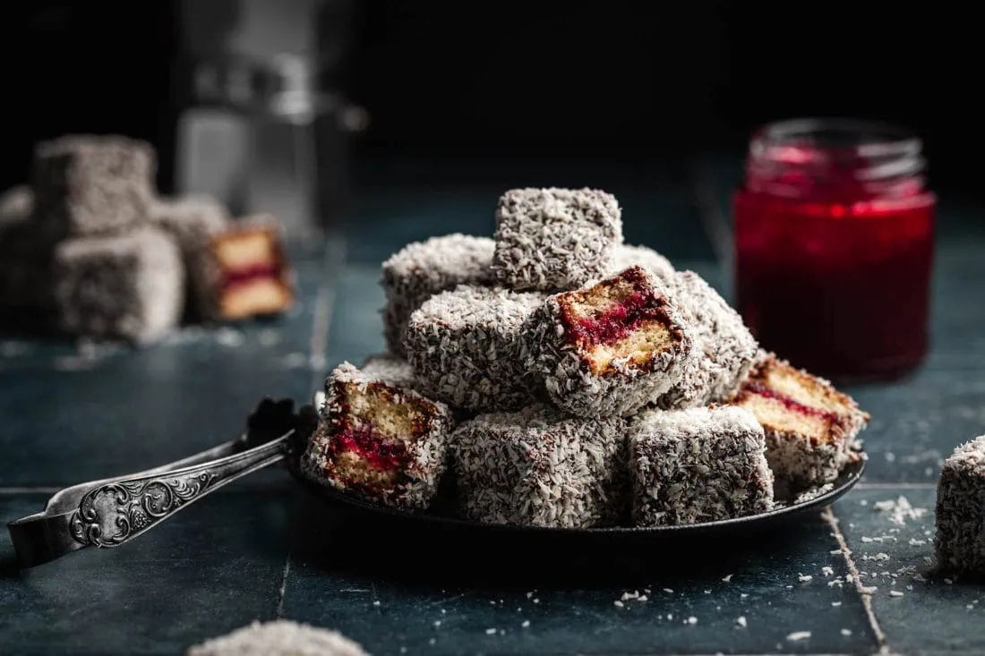 Australische Lamingtons, feine Kuchenwürfel mit Schokoladenglasur und Kokosraspeln und einer Füllung aus Marmelade auf einem Teller.