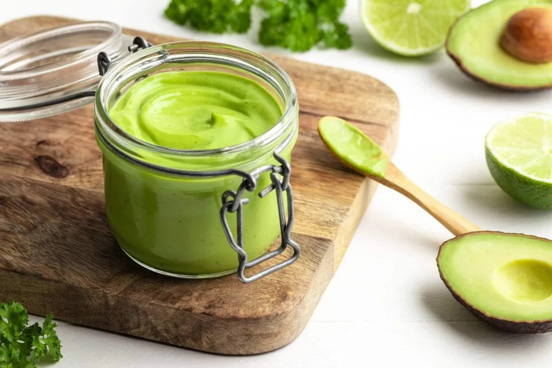 Avocado-Limetten-Mayonnaise in einem Glas. Neben dem Glas liegen ein Holzlöffel und die Zutaten für die Mayo.