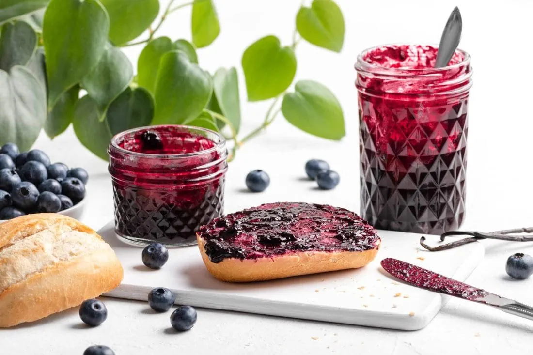 Blaubeermarmelade mit Vanille auf einem Brötchen. Daneben stehen Marmeladengläser und frische Blaubeeren.