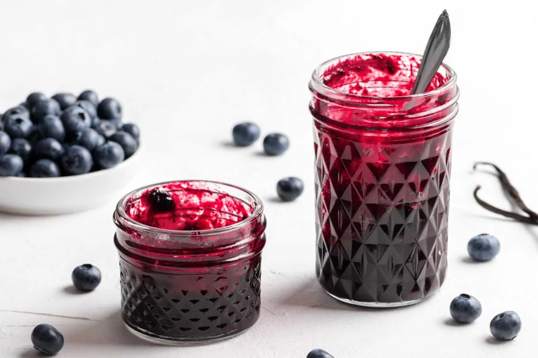 Blaubeermarmelade mit Vanille in zwei Gläsern. Daneben liegen frische Beeren.