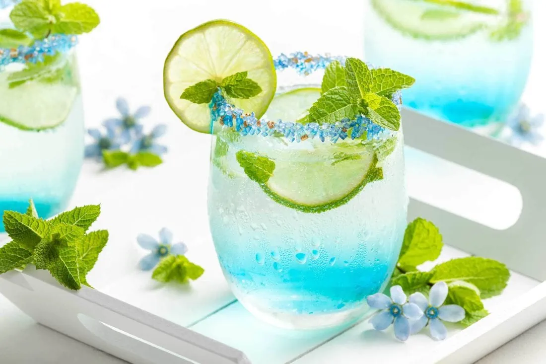 Blauer Hugo mit Blue Curaçao in einem runden Glas mit bunten Zuckerrand.