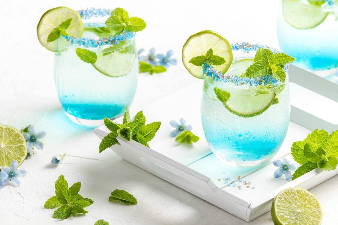 Blauer Hugo mit Blue Curaçao in drei Gläsern mit Zuckerrand und einer Garnitur aus Limette und Minze.