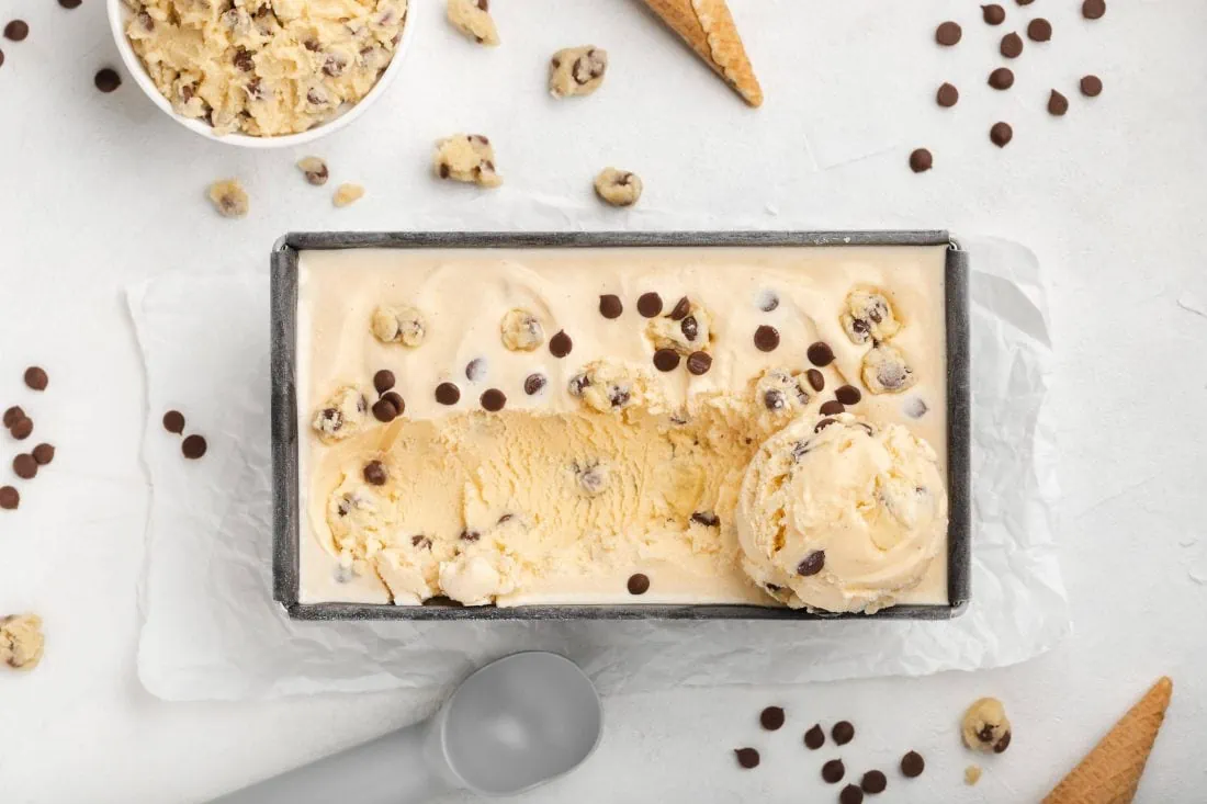 Cookie Dough Eis in einer rechteckigen Form von oben. Daneben liegen Chocolate Chips, Eiswaffeln und Teigtsücke.