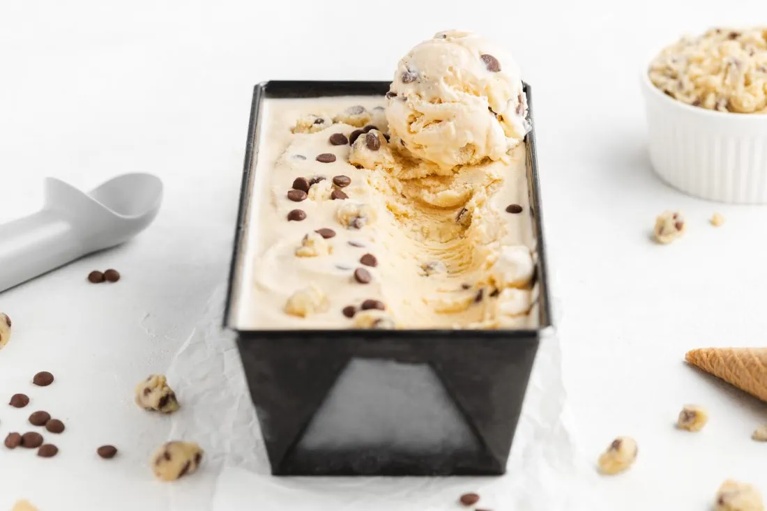 Das Cookie Dough Eis aus der Eismaschine abgefüllt in einer rechteckigen Form. Neben der Form liegt ein Eisportionierer.