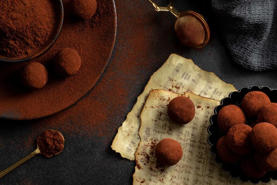 Cranberry-Lebkuchen-Pralinen neben einer Schale mit Backkakao, indem sie gewälzt werden. Daneben liegen Notenblätter mit Weihnachtsliedern.