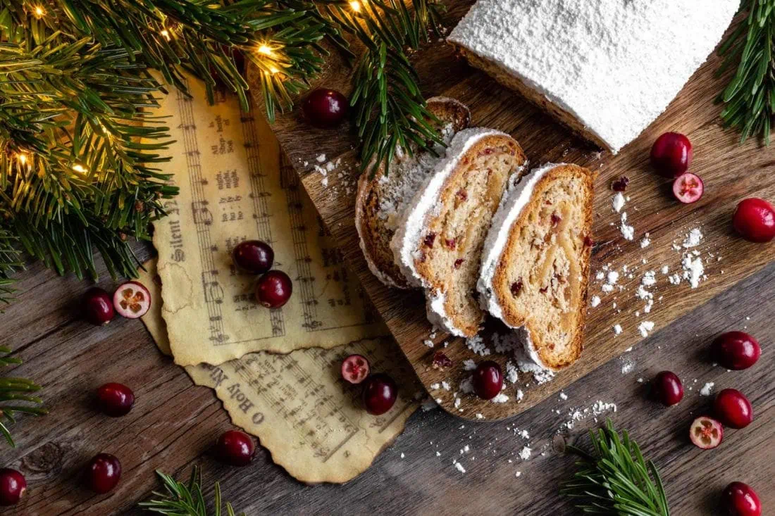Cranberry-Marzipan-Stollen in Scheiben geschnitten auf einem Holzbrett. Daneben liegen Tannenzweige und frische Cranberrys.