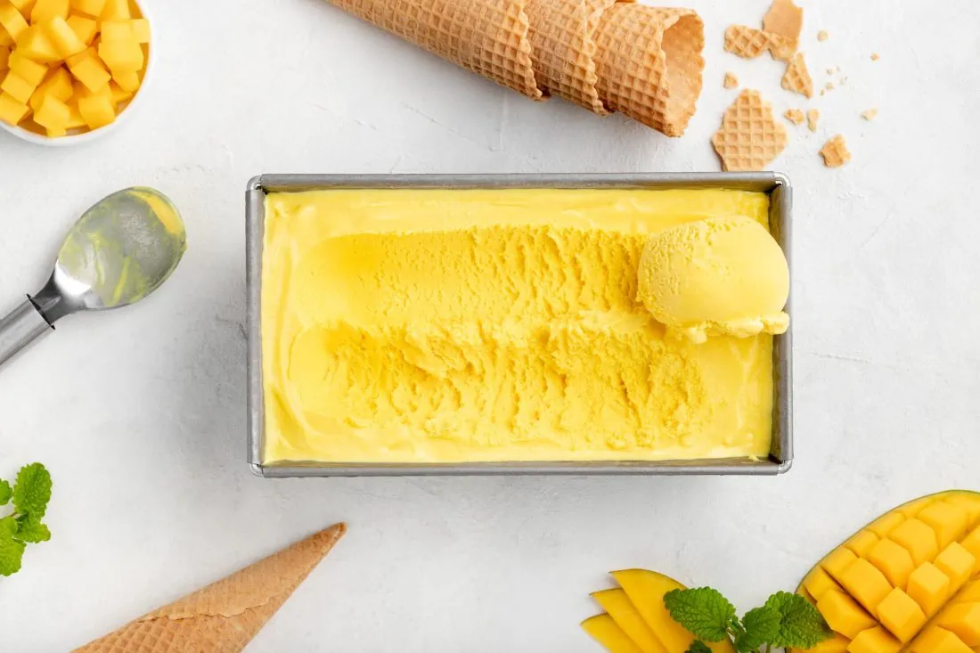 Selbstgemachtes Mangoeis aus der Eismaschine in einer rechteckigen Form. Daneben liegen frische Mangostücke und ein Eislöffel.