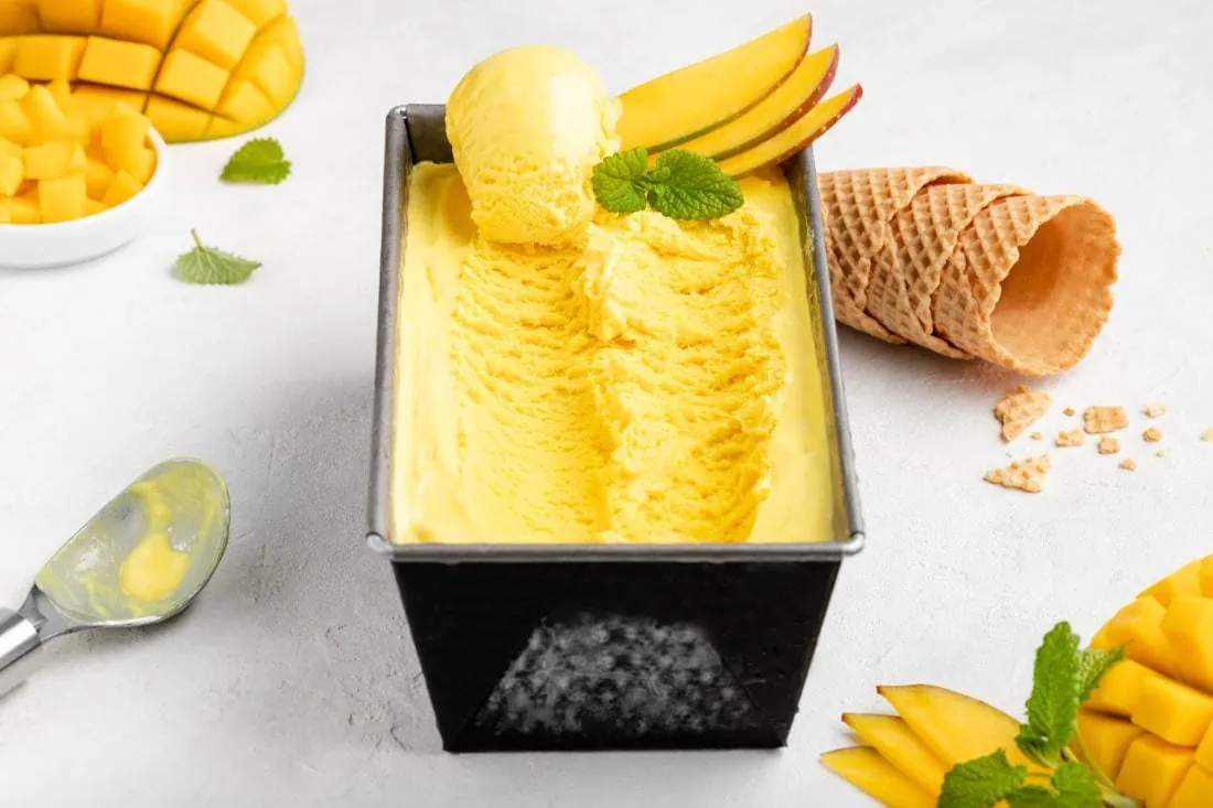 Eine rechteckige Form gefüllt mit dem cremigen Mangoeis aus der Eismaschine. Daneben liegen Eiswaffeln.