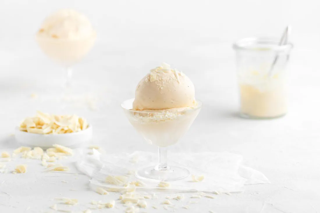 Eine Kugel cremiges, weißes Schokoladeneis aus der Eismaschine in einem Glas. Daneben liegen weiße Schokoladenraspeln.