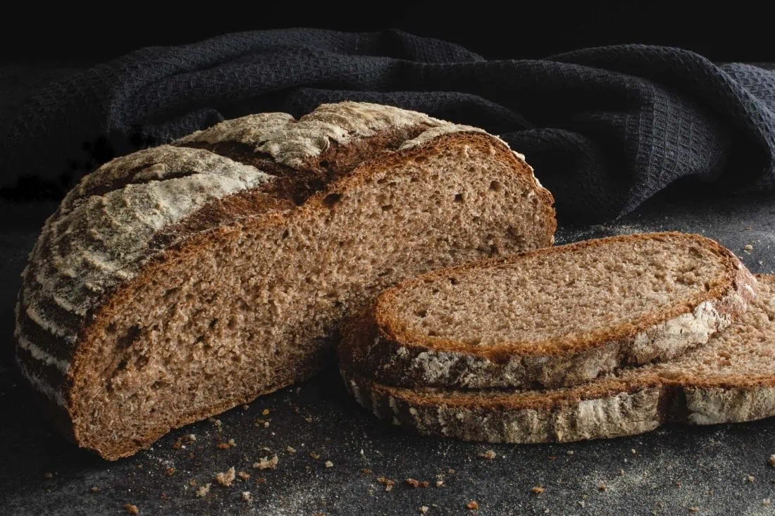 Auf einem dunklen Untergrund liegt ein aufgeschnittenes Dinkelvollkornbrot mit Sauerteig. Neben dem angeschnittenen Brotlaib liegen zwei Scheiben des Vollkornbrotes und zahlreiche Brösel.