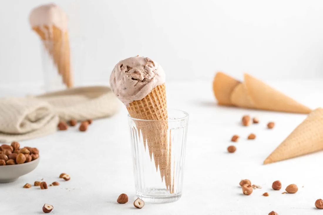 Eine Eiswaffel mit einer großen Kugel selbstgemachten Haselnusseis aus der Eismaschine. Daneben liegen weitere Eistüten und Haselnüsse.