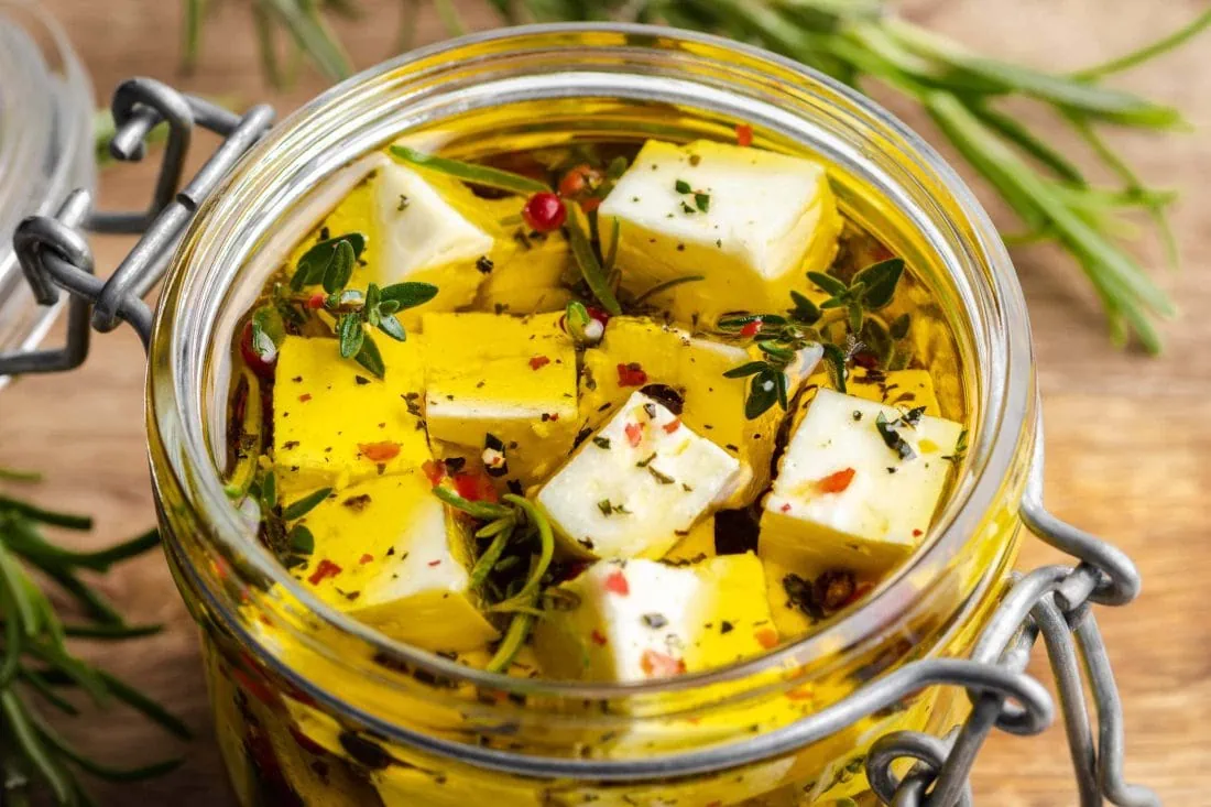 Schafskäse eingelegt in Olivenöl zusammen mit Chili, Thymian, Rosmarin und bunten Pfefferkörnern in einem Glas.